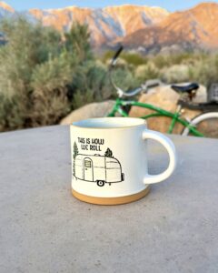 A white coffee mug with a travel trailer illustration sits on a picnic table, with a green bicycle and sunlit mountains in the background.